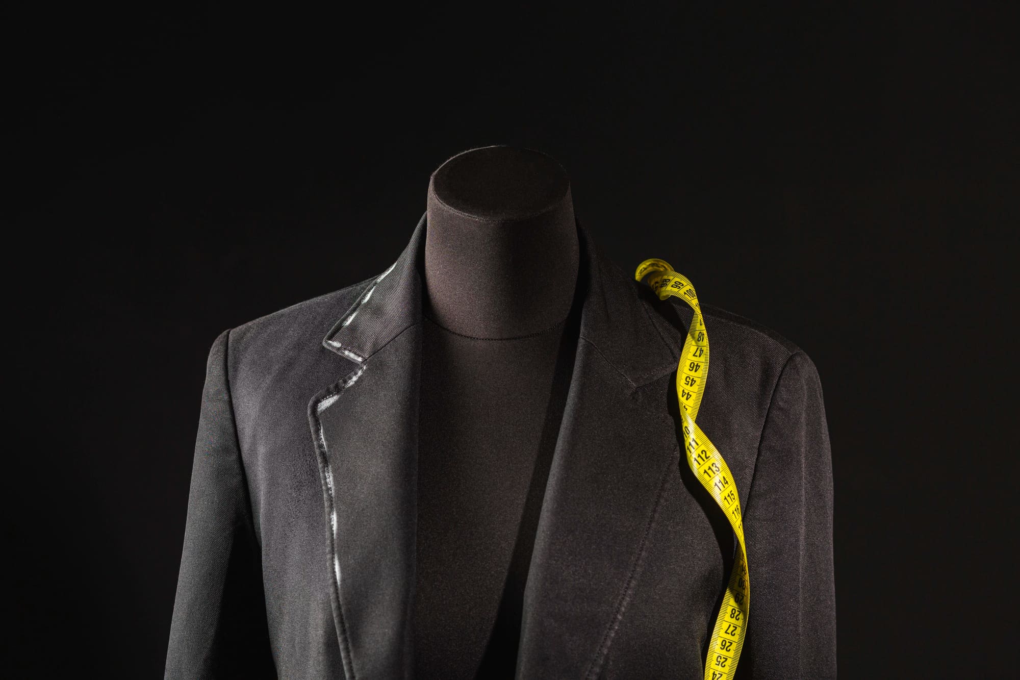 A black tailor’s mannequin dressed in a dark blazer with a yellow measuring tape draped over one shoulder, set against a black background—perfect for showcasing expert leather garment cleaning Syracuse services.