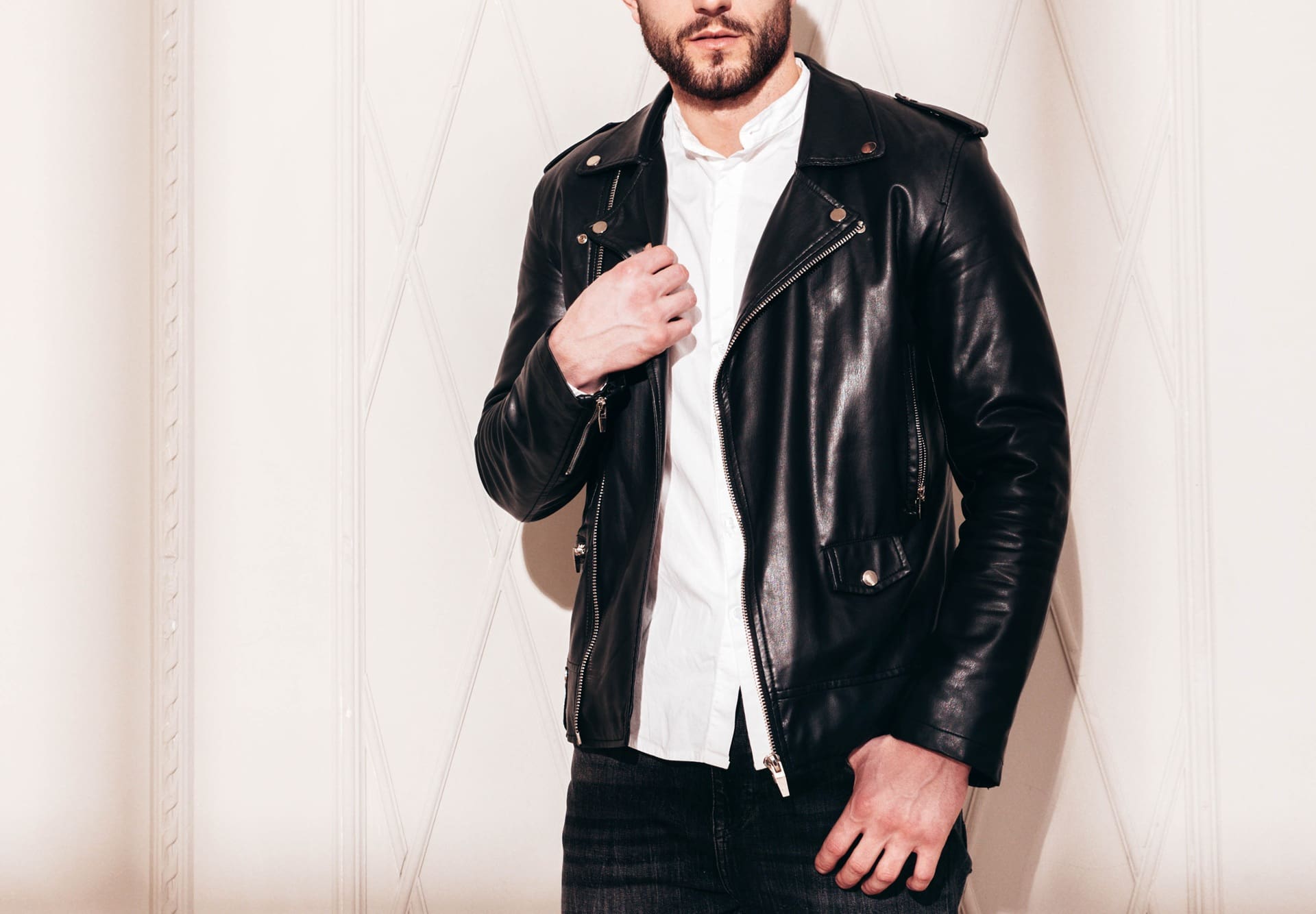 A man in a black leather jacket over a white shirt stands against a beige wall, with his left hand touching the jacket. His face is partially out of frame, much like newly cleaned gowns at Wedding dress cleaning Manlius—spotless and sharp.