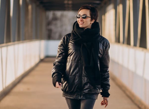 Person wearing sunglasses, a black scarf, and a black leather jacket walks across a pedestrian bridge on a sunny day, just steps away from trusted wedding dress cleaning in Baldwinsville.
