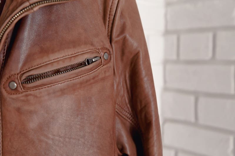 Close-up of a brown leather jacket with a visible zippered pocket, shown against a white painted brick wall background. Expert care like wedding dress cleaning in Syracuse ensures lasting quality for your cherished pieces.
