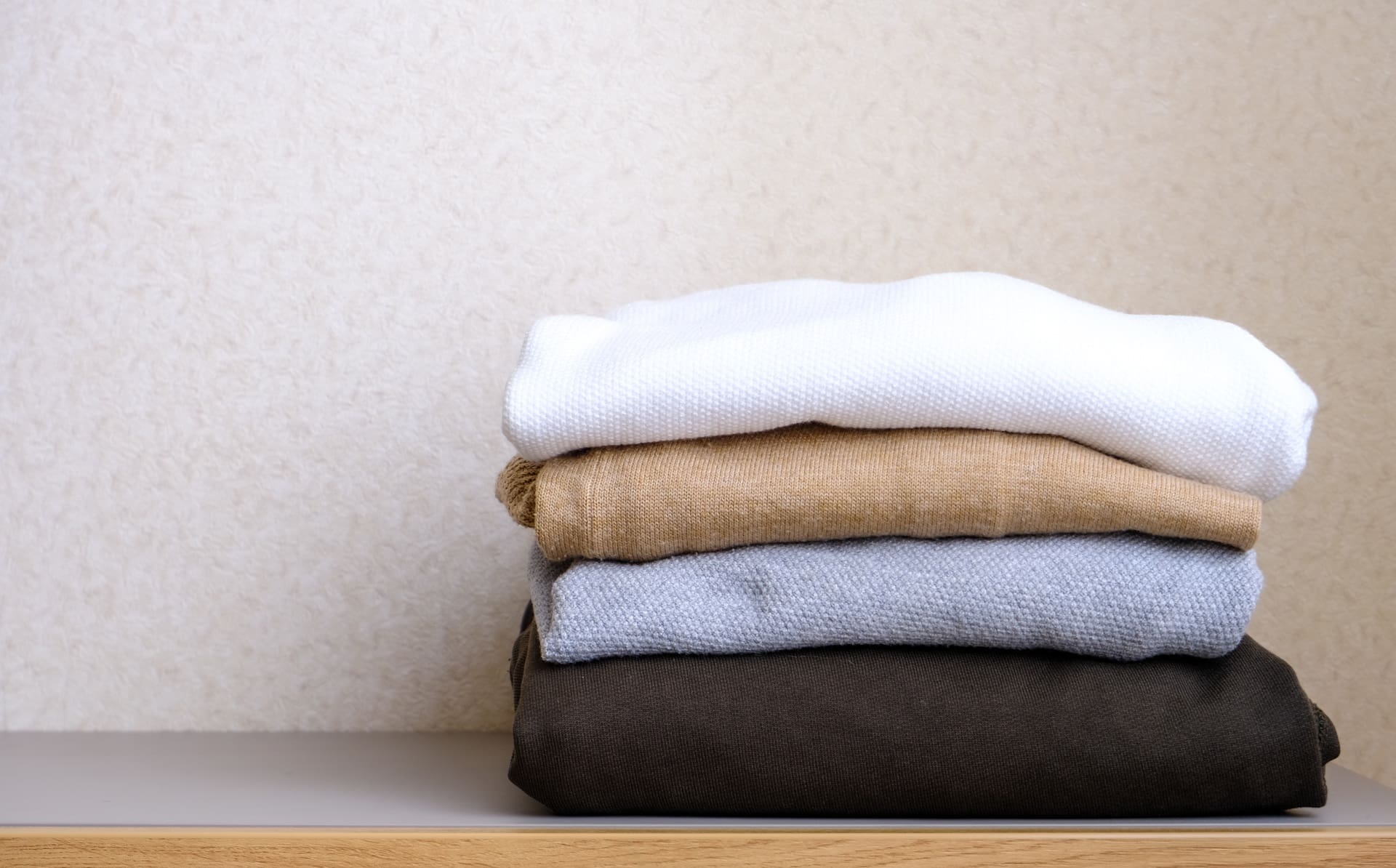 A neatly folded stack of four sweaters in white, beige, gray, and dark brown colors sits on a wooden surface against a light-colored wall.