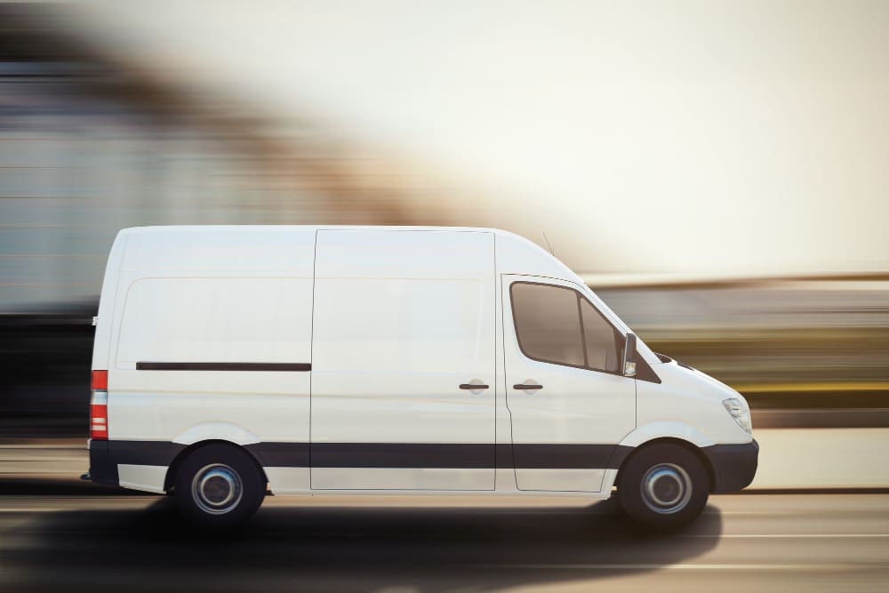 A white cargo van drives quickly on a road with a blurred background, suggesting motion.