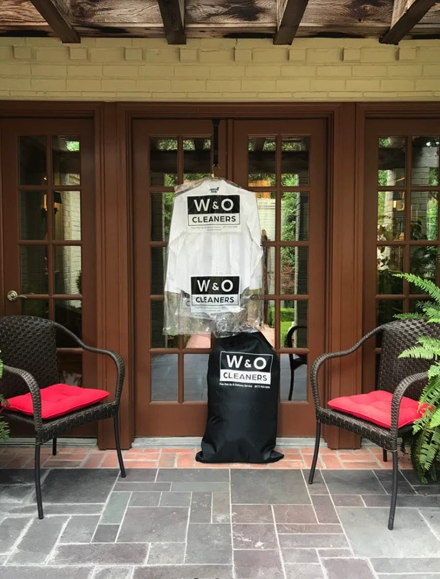 A dry cleaning delivery with clothing in plastic covers and a black W&O Cleaners bag is placed in front of brown double doors between two wicker chairs with red cushions.