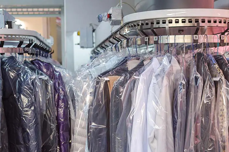 Clothes covered in plastic hang on a rotating conveyor rack in a dry cleaning facility.