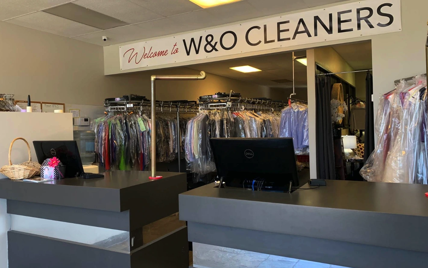 Interior of a dry cleaning shop with clothes on racks behind two counters, a computer, a basket, and a "Welcome to W&O Cleaners" sign overhead.