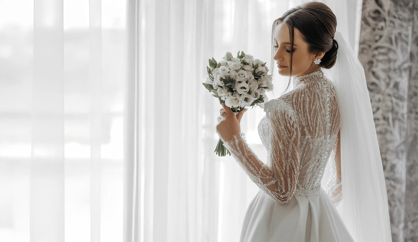 A bride in a detailed lace wedding dress holds a bouquet of white flowers and stands beside sheer curtains, looking down.