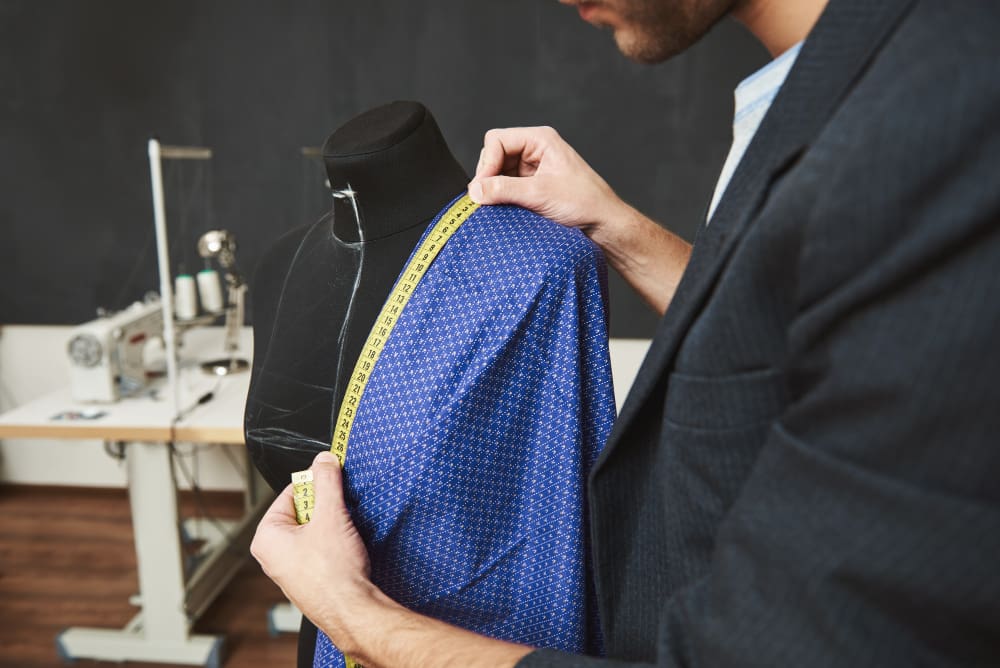 A tailor measures fabric draped over a dress form using a measuring tape in a sewing studio.