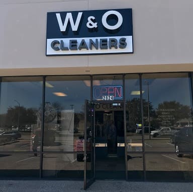 Storefront of W & O Cleaners with an illuminated "OPEN" sign above the door, glass windows, and reflections of cars in the parking lot.