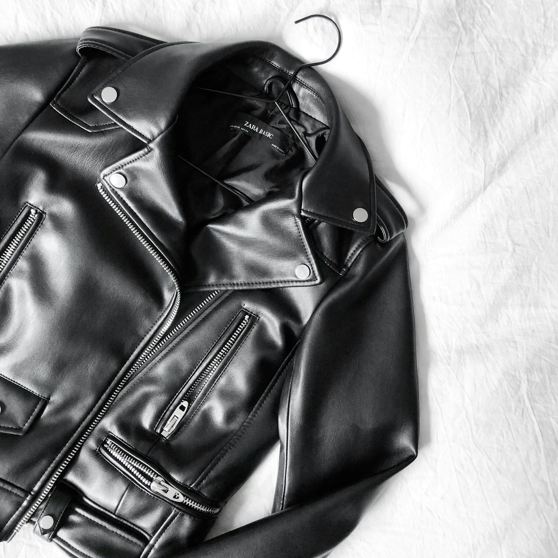 A black faux leather biker jacket with silver zippers and buttons is displayed on a hanger against a white background.