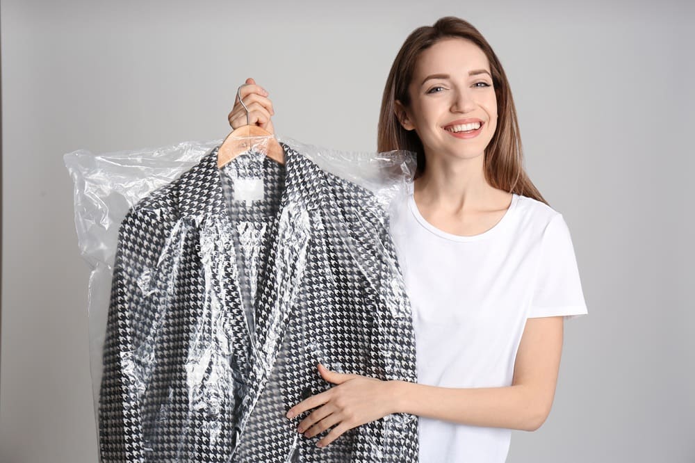 A woman in a white shirt smiles while holding a houndstooth-patterned jacket covered in clear plastic on a hanger.