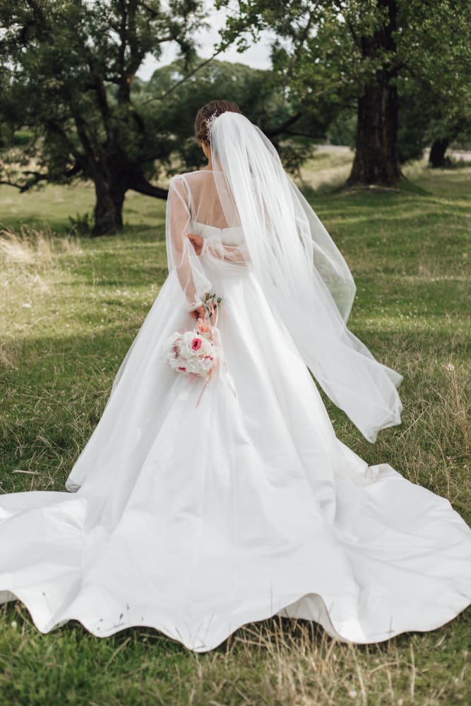 A bride in a white wedding dress and veil stands outdoors on grass, facing away, holding a bouquet of pink and white flowers.