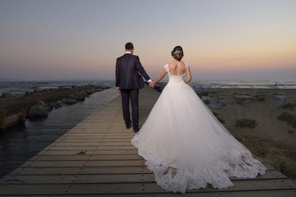 A bride and groom in formal wedding attire walk hand in hand on a wooden pier at sunset, facing the sea.