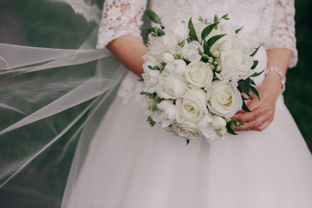 A bride in a white wedding dress holding a bouquet of white roses and greenery, with a lace veil draped to the side.