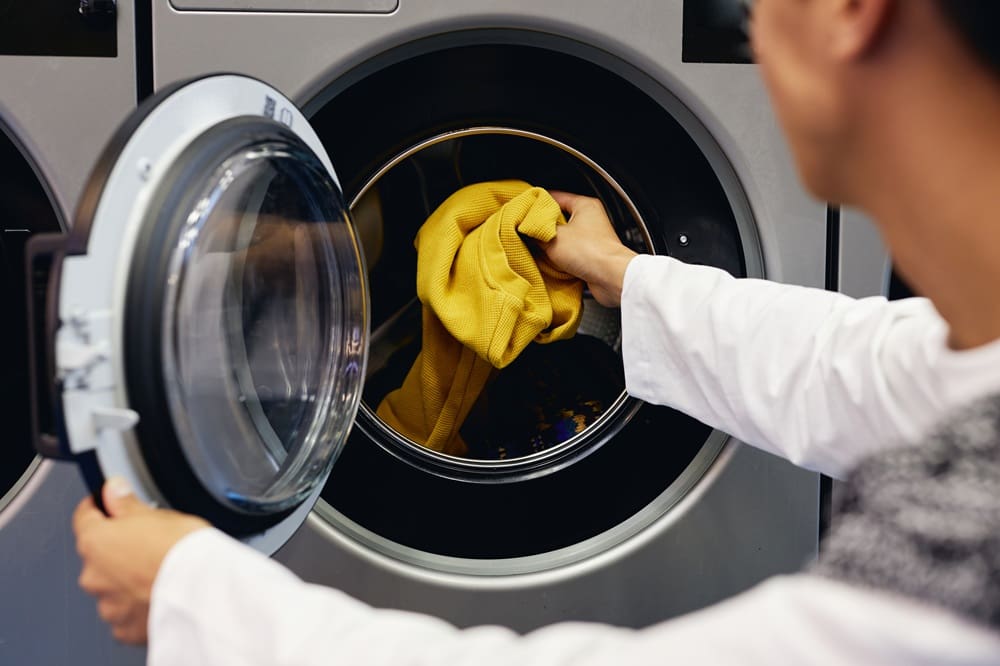 A person places a yellow garment into the open door of a front-loading washing machine.