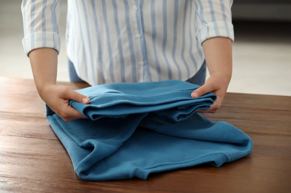 Person folding a blue sweatshirt on a wooden table.