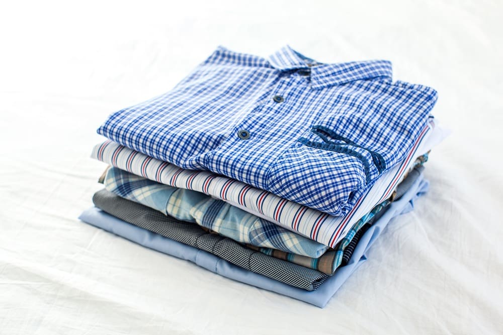 A neatly folded stack of men's shirts in various patterns and colors, placed on a white surface.