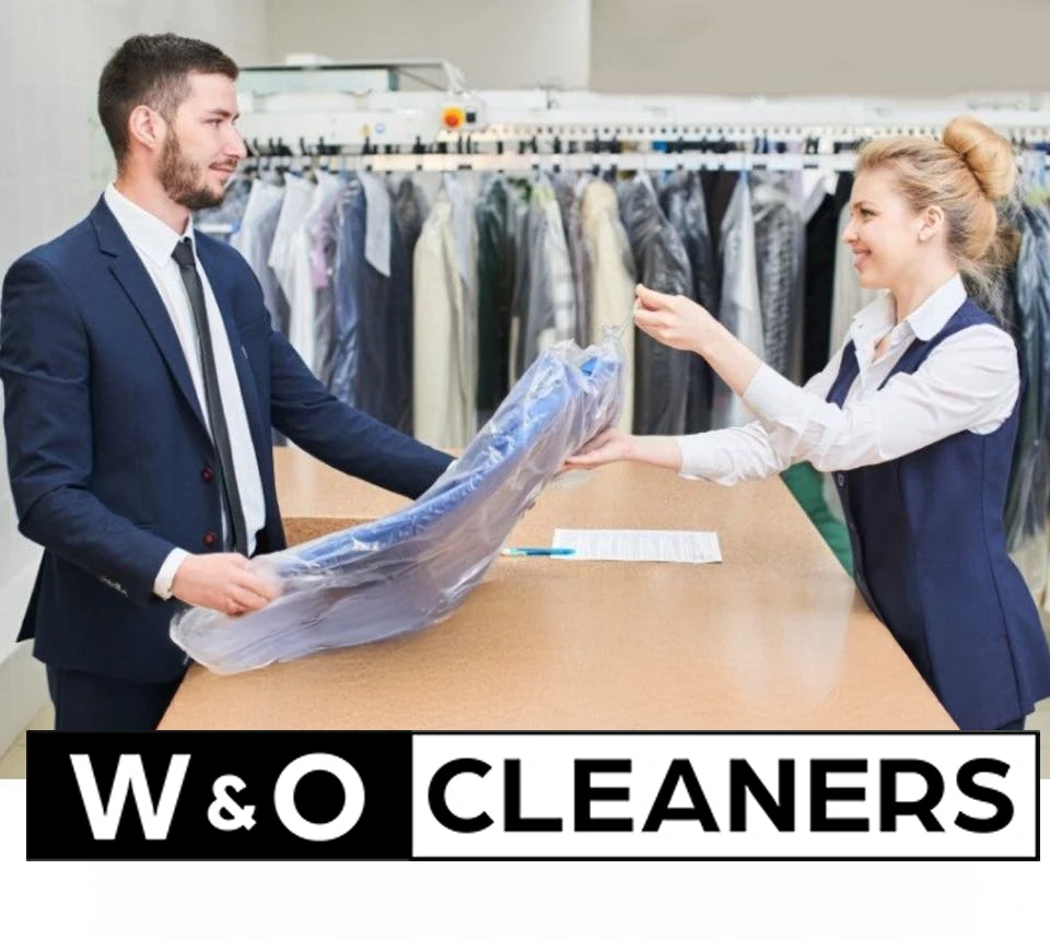 A man in a suit receives dry-cleaned clothes from a woman behind a counter at a dry cleaning shop. 